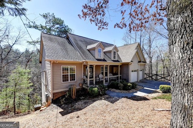 cape cod home featuring driveway, roof with shingles, an attached garage, covered porch, and fence