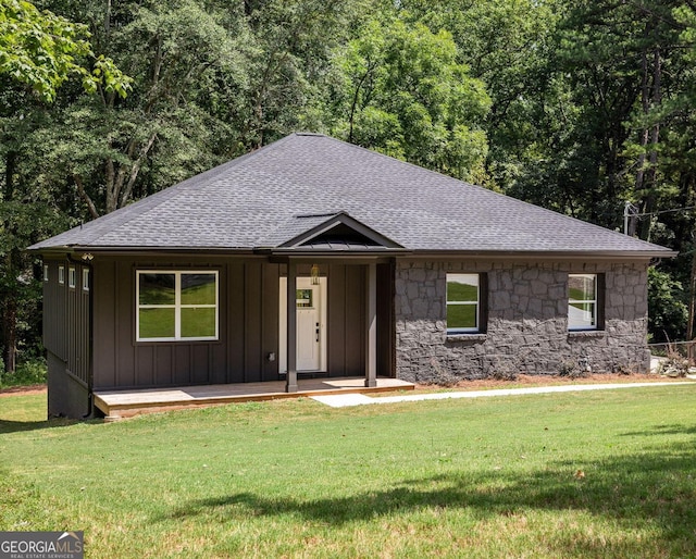 view of front facade featuring a shingled roof, a front lawn, stone siding, a view of trees, and board and batten siding