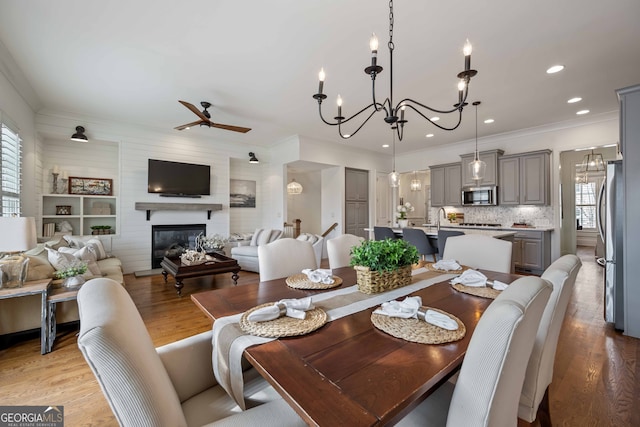 dining room featuring light wood-style flooring, ceiling fan with notable chandelier, a glass covered fireplace, recessed lighting, and crown molding