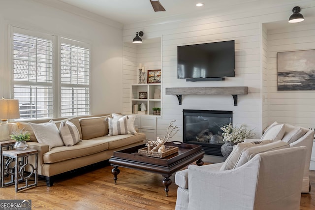 living area featuring wooden walls, a ceiling fan, wood finished floors, crown molding, and a large fireplace