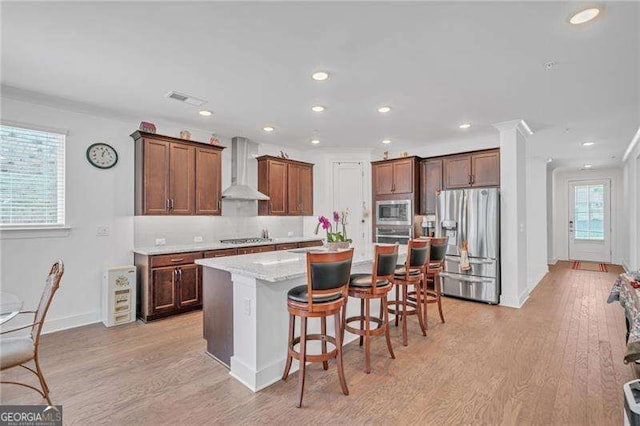 kitchen with wall chimney range hood, a kitchen island, appliances with stainless steel finishes, and light wood-style floors