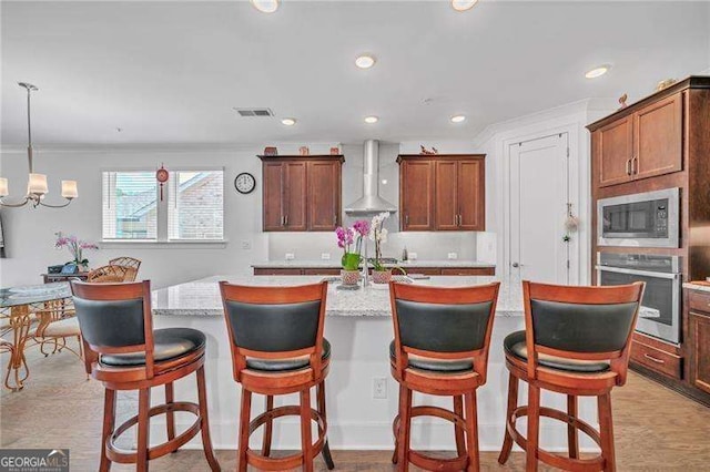 kitchen with wall chimney exhaust hood, appliances with stainless steel finishes, an inviting chandelier, a kitchen island with sink, and recessed lighting