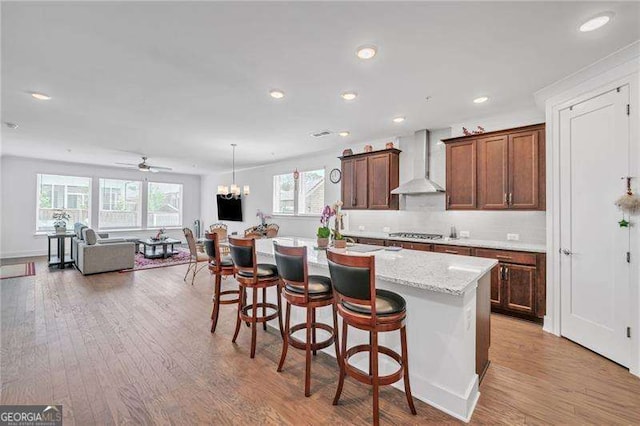 kitchen featuring a center island with sink, wall chimney exhaust hood, a breakfast bar, light wood-type flooring, and stainless steel gas cooktop