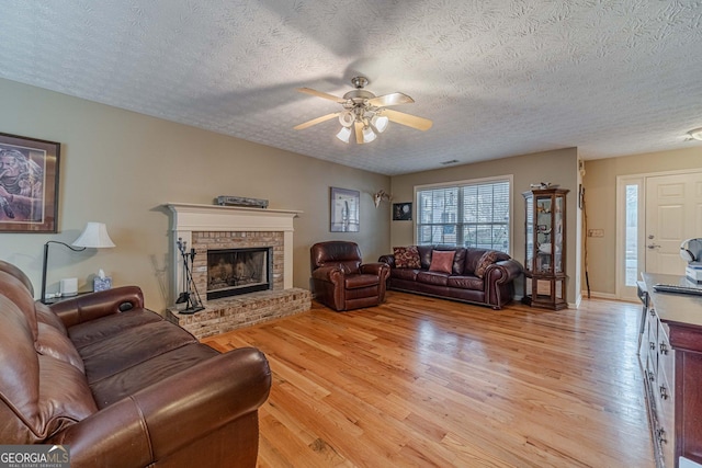 living room with visible vents, a brick fireplace, light wood-type flooring, a textured ceiling, and a ceiling fan