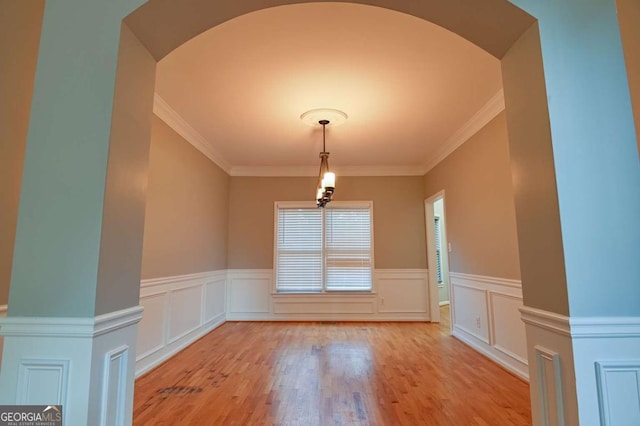 unfurnished dining area featuring a wainscoted wall, arched walkways, light wood-style flooring, and crown molding