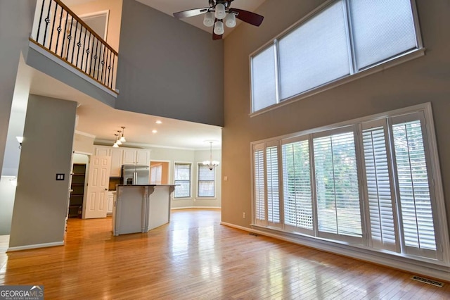 unfurnished living room featuring visible vents, baseboards, light wood finished floors, a high ceiling, and ceiling fan with notable chandelier