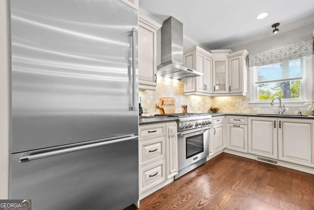 kitchen with premium appliances, dark wood-style floors, dark countertops, a sink, and wall chimney exhaust hood