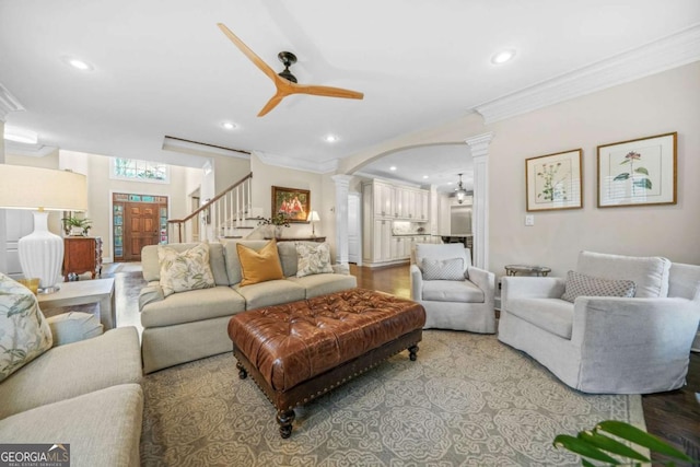 living area with arched walkways, a ceiling fan, stairs, crown molding, and ornate columns