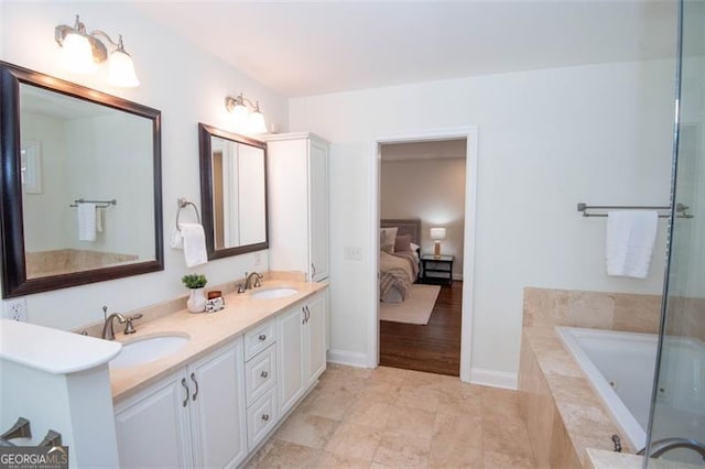 ensuite bathroom featuring double vanity, tiled bath, a sink, and baseboards