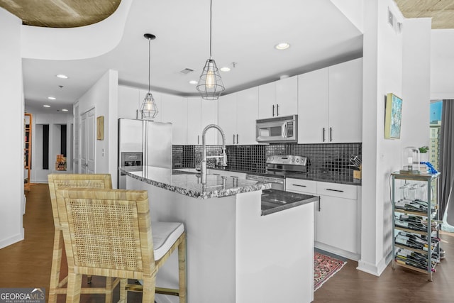 kitchen with a center island with sink, stainless steel appliances, decorative backsplash, dark wood-type flooring, and decorative light fixtures