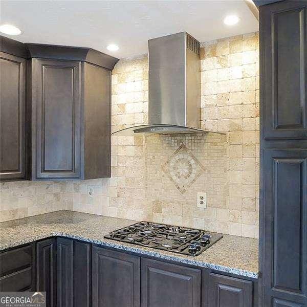 kitchen with dark brown cabinetry, light stone countertops, wall chimney range hood, gas stovetop, and backsplash