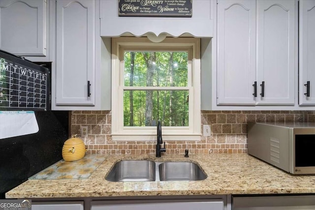 kitchen with stainless steel microwave, decorative backsplash, white cabinetry, a sink, and light stone countertops