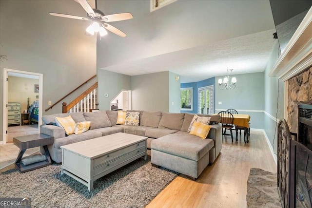 living room with baseboards, stairway, a stone fireplace, light wood-type flooring, and ceiling fan with notable chandelier