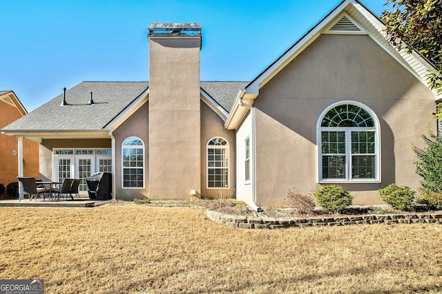 rear view of house with stucco siding, a lawn, a patio, a shingled roof, and a chimney