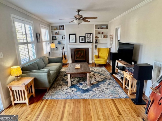 living room with a fireplace with flush hearth, ceiling fan, wood finished floors, crown molding, and built in shelves