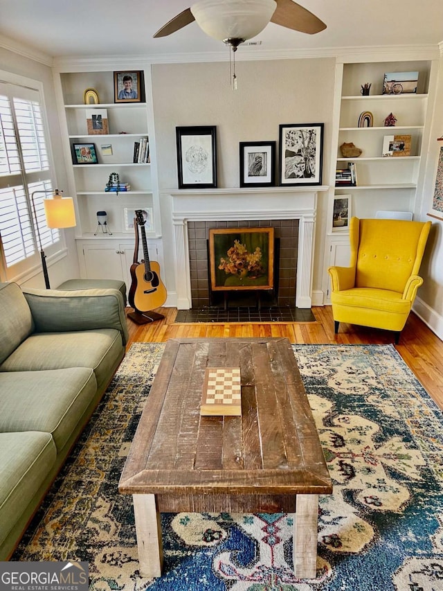 living area featuring ceiling fan, a tile fireplace, wood finished floors, built in features, and ornamental molding