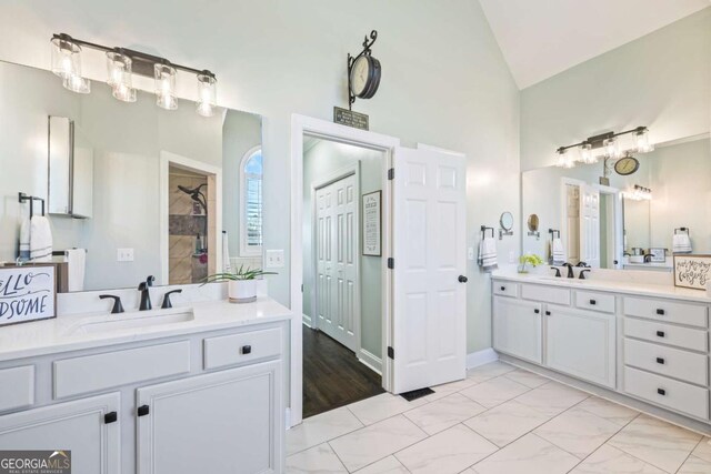 bathroom featuring lofted ceiling, tiled shower, two vanities, and a sink