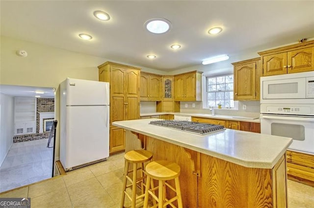 kitchen with light countertops, a brick fireplace, a kitchen island, white appliances, and a kitchen bar