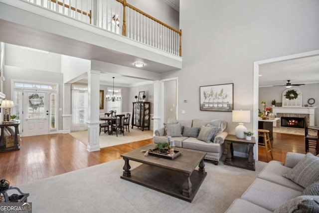 living area featuring wood finished floors, decorative columns, a towering ceiling, a brick fireplace, and ceiling fan with notable chandelier