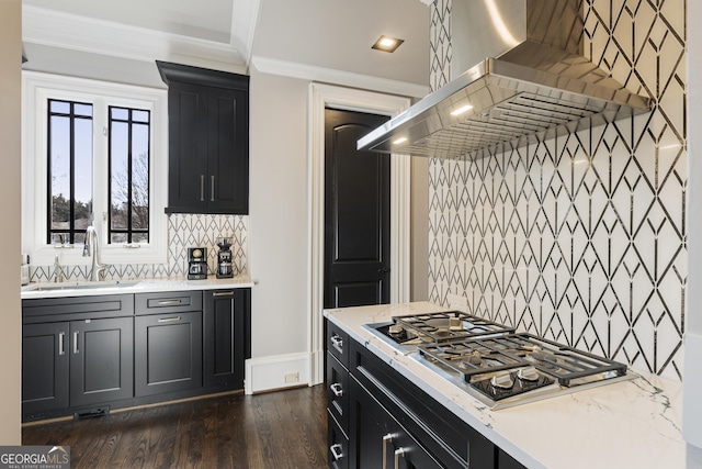 kitchen with stainless steel gas cooktop, a sink, crown molding, wall chimney range hood, and dark cabinets