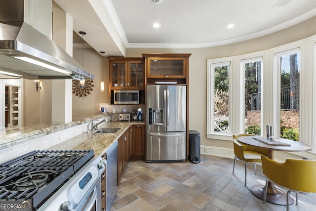 kitchen featuring baseboards, wall chimney range hood, appliances with stainless steel finishes, brown cabinetry, and a sink