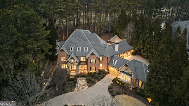 view of front facade with decorative driveway, stone siding, a wooded view, and a chimney