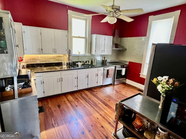 kitchen featuring white cabinets, dark countertops, wall chimney exhaust hood, stainless steel appliances, and a sink