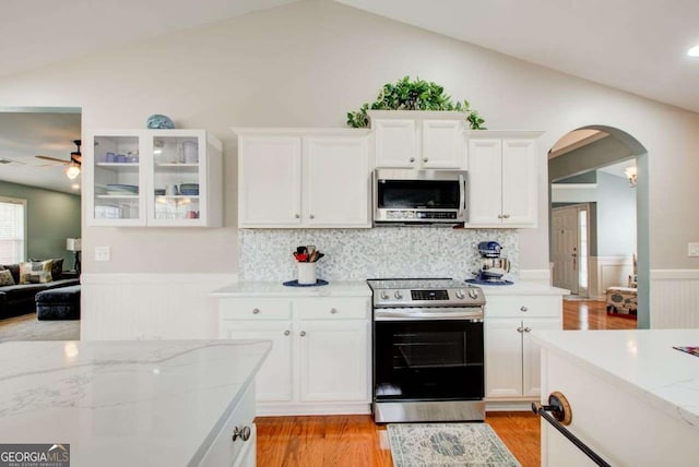 kitchen with a wainscoted wall, lofted ceiling, arched walkways, white cabinets, and appliances with stainless steel finishes