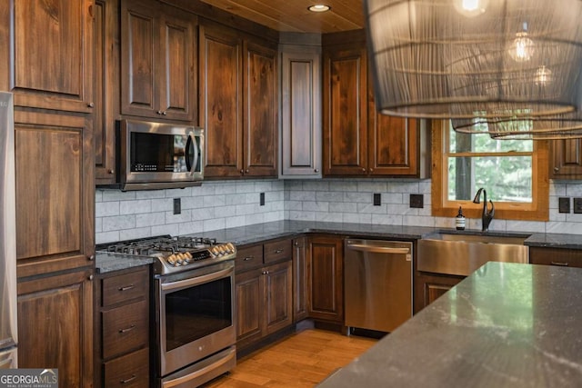 kitchen featuring stainless steel appliances, a sink, light wood-style floors, backsplash, and dark stone counters