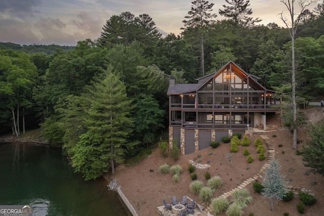 back of house at dusk featuring an attached garage, driveway, stairway, a chimney, and a wooded view