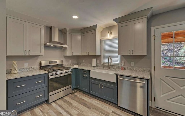 kitchen featuring a sink, light wood-style floors, appliances with stainless steel finishes, wall chimney range hood, and light stone countertops