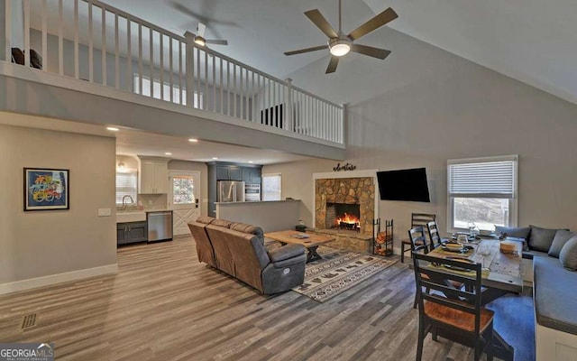 living room featuring light wood-type flooring, high vaulted ceiling, a ceiling fan, a stone fireplace, and baseboards