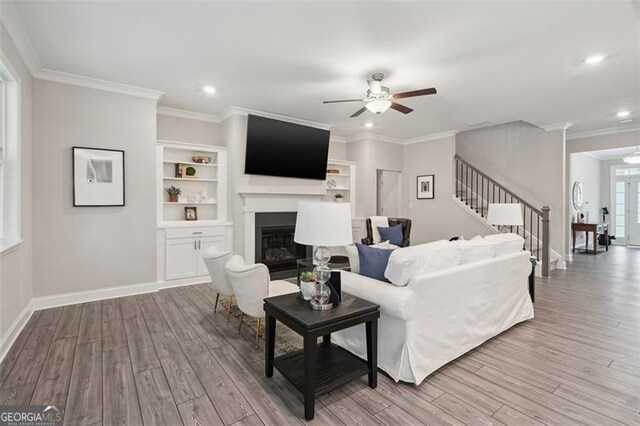 living area featuring stairs, ornamental molding, a glass covered fireplace, and light wood-style flooring