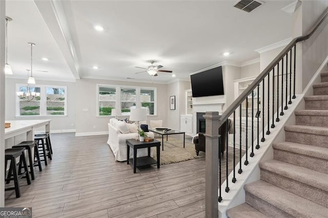 living room featuring crown molding, ceiling fan with notable chandelier, visible vents, wood finished floors, and stairs