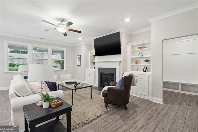 living area featuring ornamental molding, a glass covered fireplace, ceiling fan, wood finished floors, and baseboards