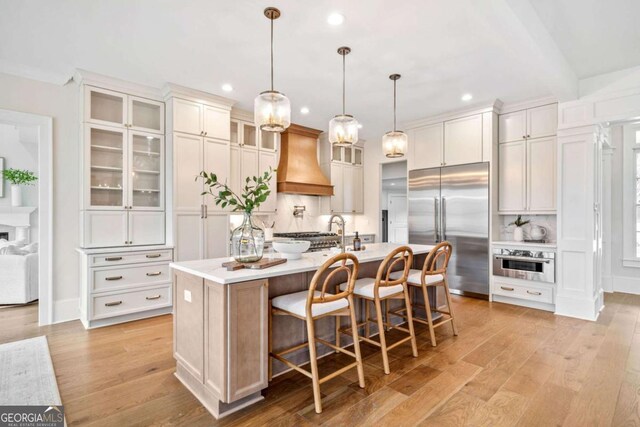 kitchen featuring an island with sink, custom range hood, appliances with stainless steel finishes, light countertops, and light wood-type flooring