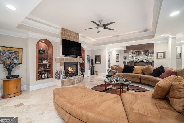 living area featuring a tray ceiling, decorative columns, ceiling fan, a tiled fireplace, and crown molding