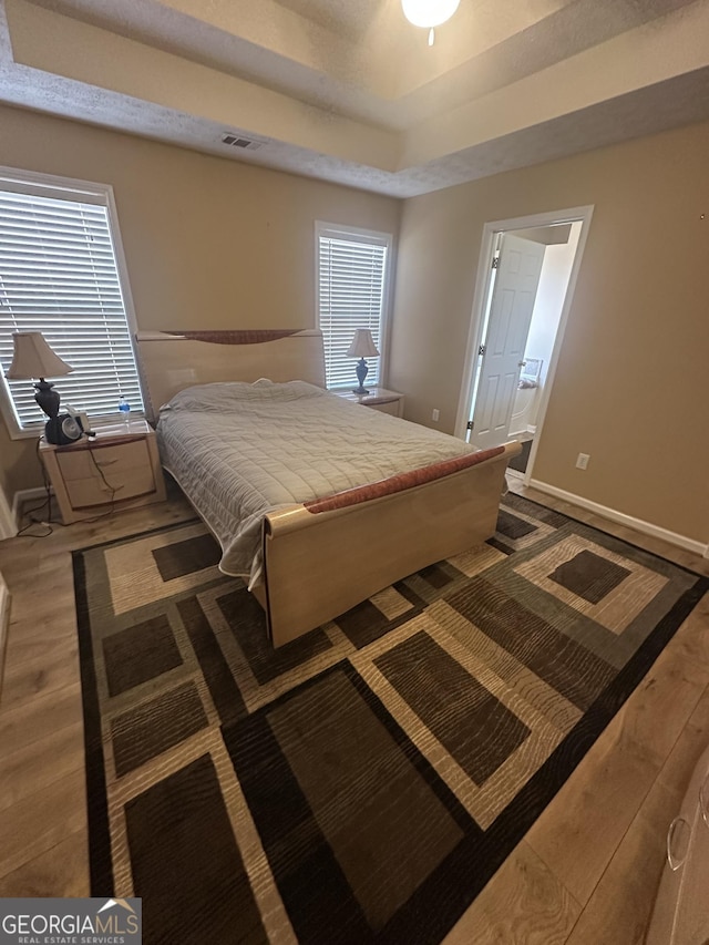 bedroom featuring visible vents, baseboards, a raised ceiling, ensuite bath, and wood finished floors