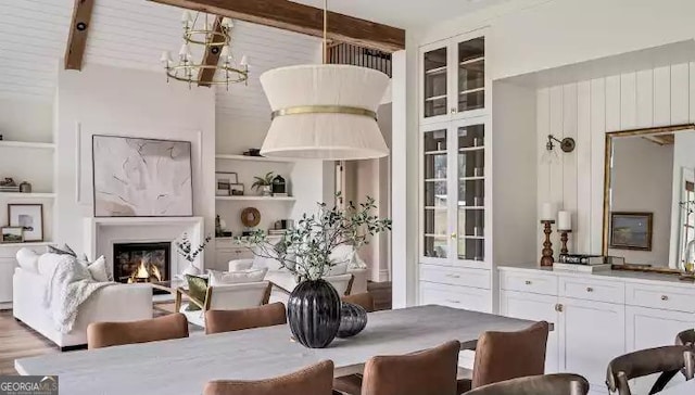 dining room featuring built in shelves, beamed ceiling, a lit fireplace, and an inviting chandelier