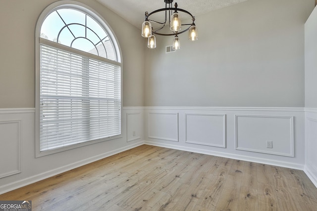 unfurnished dining area with light wood-style flooring, a decorative wall, a notable chandelier, visible vents, and wainscoting