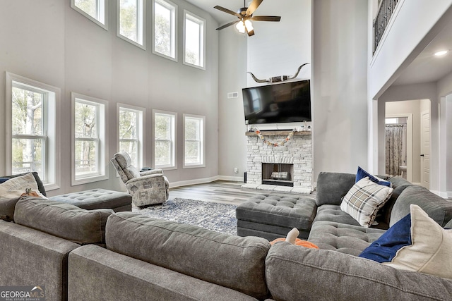 living area with baseboards, a ceiling fan, a towering ceiling, wood finished floors, and a stone fireplace