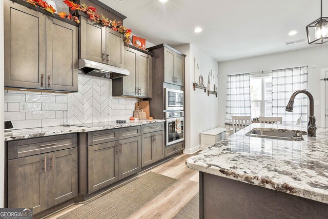 kitchen featuring light stone counters, light wood-style flooring, under cabinet range hood, a sink, and appliances with stainless steel finishes