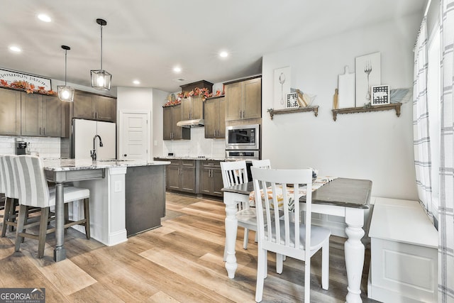 kitchen featuring stainless steel appliances, light wood-type flooring, decorative backsplash, an island with sink, and decorative light fixtures