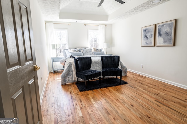 bedroom with a textured ceiling, ceiling fan, baseboards, light wood-style floors, and a raised ceiling