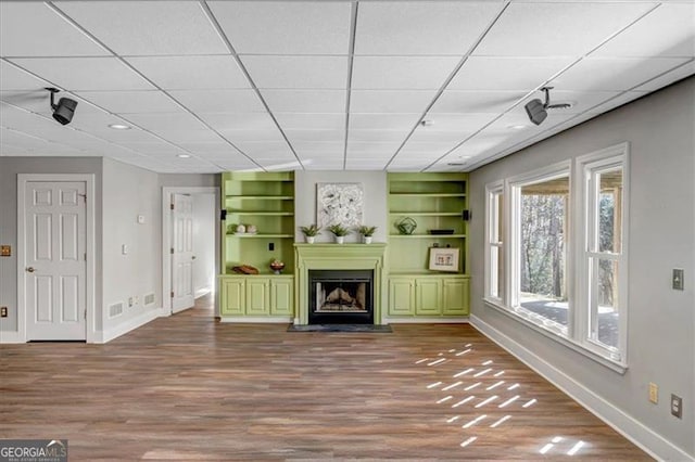 unfurnished living room featuring built in shelves, wood finished floors, baseboards, a fireplace, and a paneled ceiling