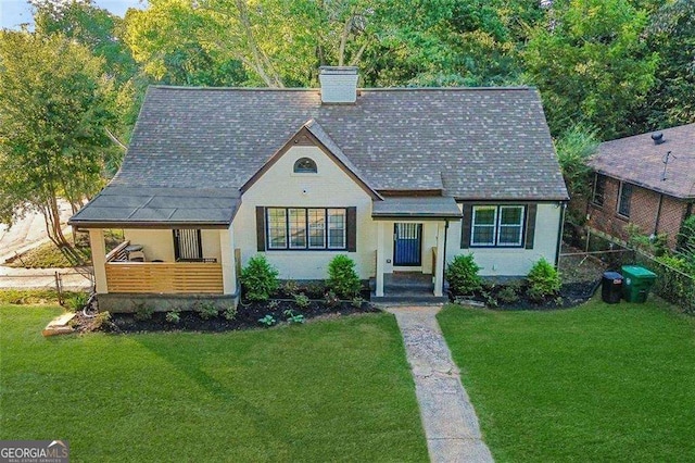 tudor-style house with a shingled roof, a front lawn, fence, and a chimney