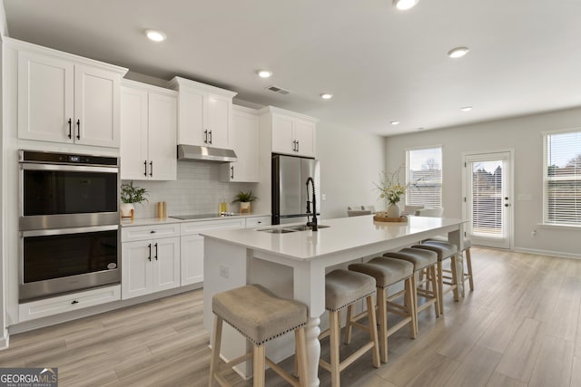 kitchen with a center island with sink, backsplash, stainless steel appliances, under cabinet range hood, and a sink