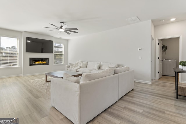 living area with ceiling fan, a glass covered fireplace, light wood-style flooring, and baseboards