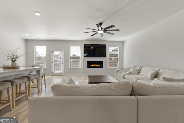 living room featuring light wood finished floors, a glass covered fireplace, and a ceiling fan