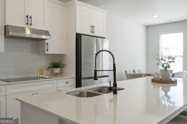 kitchen with black electric stovetop, tasteful backsplash, a sink, an island with sink, and under cabinet range hood